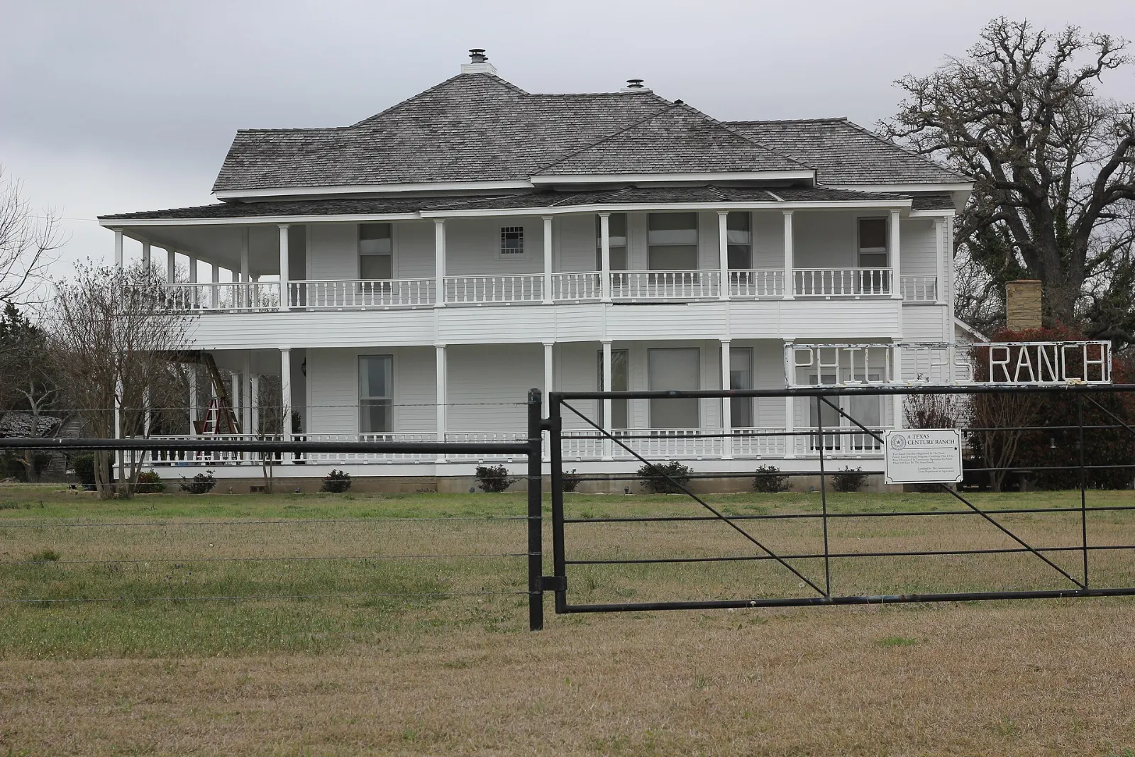A white two-story ranch house with wraparound porches behind a pipe gate